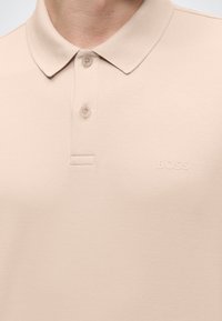 Polo beige avec col boutonné et logo "BOSS" discret sur la poitrine, porté par une personne à la peau claire.