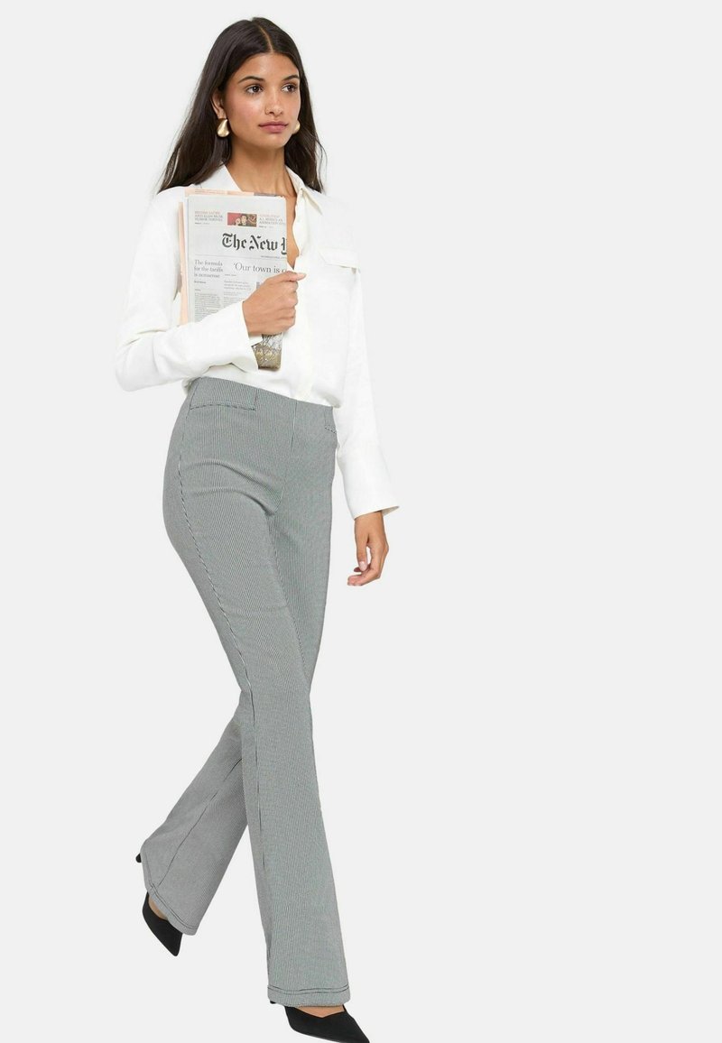 Chemise blanche à manches longues, pantalon gris à motif avec jambes évasées et talons noirs. Modèle tenant un journal, coiffure est longue et lisse.