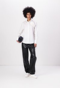 Camicia bianca con bottoni e decorazioni floreali, abbinata a pantaloni in ecopelle neri e sneakers bianche; tiene una clutch scura.