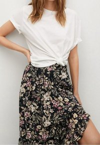T-shirt blanc à manches courtes noué à la taille, associé à une jupe longue noire à fleurs avec des motifs roses et crème et un tissu texturé.