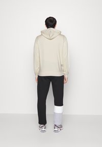 Sweat à capuche beige clair avec une texture douce, associé à un pantalon de jogging noir avec un ourlet à blocs de couleurs blanc et gris.