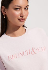 Svijetlo ružičasta majica s grafikonom "BRUNCH & YAP" u tamnijoj ružičastoj boji. Mekana, glatka tekstura s dizajnom okruglog vrata. Minimalistički stil.