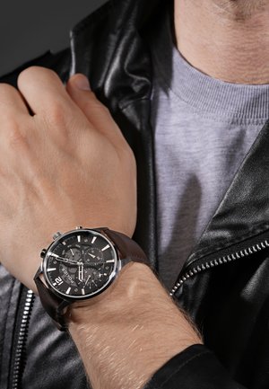 Orologio con cinturino in pelle marrone, quadrante nero, dettagli in argento e funzionalità cronografo. Indossato su un polso, accompagnato da una giacca in pelle nera.