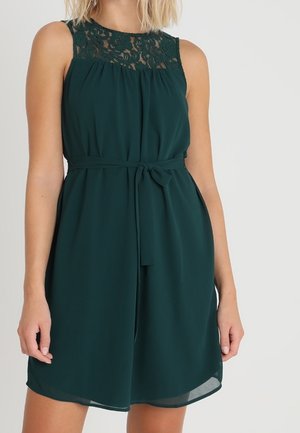 Sleeveless dark green dress with lace yoke and waist tie, worn by a person with shoulder-length blonde hair against a plain light background.
