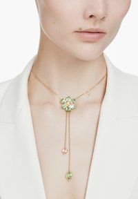 Delikat guldhalskæde med grøn og pink blomstervedhæng af ædelsten og to hængende kæder, båret med hvid blazer og bar bryst.