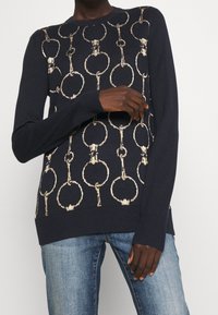 Tmavě modrý svetr s motivem zlatých řetězových oček. Vyrobený z měkké látky, má kulatý výstřih a žebrované manžety a lem.