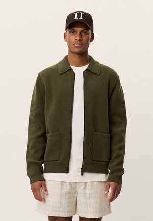 Giacca a maglia verde oliva con zip frontale, colletto a punta, due tasche frontali, indossata sopra una t-shirt bianca e pantaloncini crema con trama.