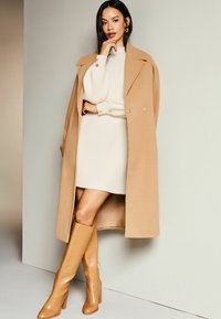 Béžový vlnený kabát s veľkými klopami, skombinovaný so smotanovým rebrovaným šatami. Modelka má na sebe svetlohnedé nadkolenné stiletto čižmy.
