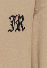 Pull beige avec un tricot texturé, arborant un logo noir brodé en évidence sur le devant. Matériau lisse et doux présenté en détail.