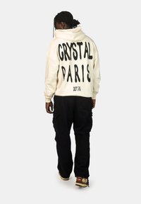 Felpa crema con testo nero audace "CRYSTAL PARIS" sul retro, abbinata a pantaloni cargo neri e sneakers multicolori.
