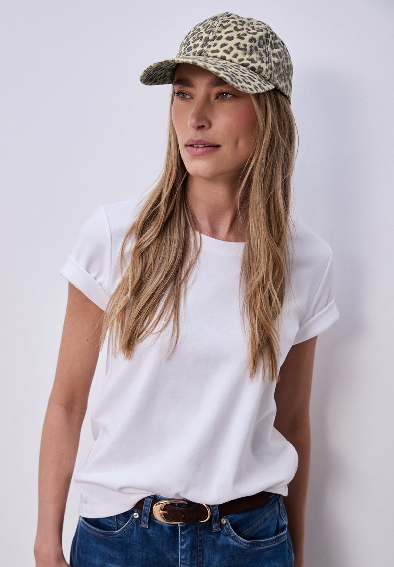 Femme aux longs cheveux blonds portant un t-shirt blanc à manches retroussées, une casquette à imprimé léopard et un jean bleu avec une ceinture marron, debout contre un mur blanc.
