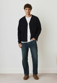 Marinblå kofta med bruna knappar, bärs över en vit skjorta, tillsammans med mörkblå jeans och bruna skor, stående mot en neutral bakgrund.