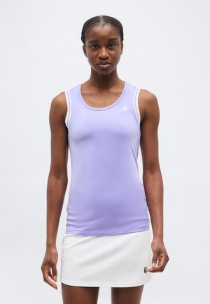 Moteris dėvi rankovių neturintį levandų atspalvio sportinį marškinėlį ir baltą sijoną, stovinti priešais paprastą šviesų foną.