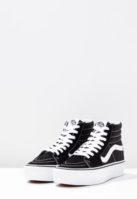 Un paio di sneakers Vans nere alte con lacci bianchi, striscia laterale bianca e suole di gomma bianca spessa su una superficie bianca.