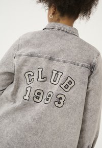 Grå denimjakke med rygtryk af "CLUB 1993" i sort. Designet inkluderer en klassisk krave og knapmanchetter.
