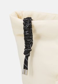 Sac de couleur crème avec une texture douce et gonflée. Doté d'une sangle noire tressée avec des accents blancs et des extrémités métalliques pour plus de détails.