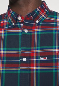 Camisa de cuadros abotonada con una mezcla de líneas rojas, verdes, azules y amarillas sobre un fondo azul marino. Incluye botones blancos y un pequeño logo.