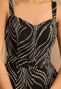 Fekete ruha, amely testre szabott felsőrészből, széles pántokból, szív alakú kivágásból és fehér pöttyös mintából áll; tartalmaz egy hozzáillő övet fém kiegészítőkkel.