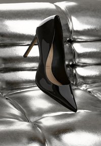 Chaussure à talon haut en cuir verni noir avec un bout pointu et un talon aiguille, sur fond argenté brillant. Surface lisse, design minimaliste.