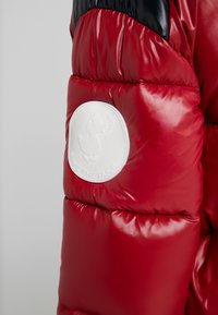 Manche de doudoune brillante rouge et noire arborant un patch circulaire blanc avec un logo de canard et le texte "SAVE THE DUCK".