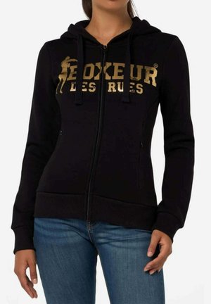 Frau trägt einen schwarzen Kapuzenpullover mit Reißverschluss und goldfarbenen "BOXEUR DES RUES" Schriftzug sowie Silhouette, kombiniert mit blauen Jeans.