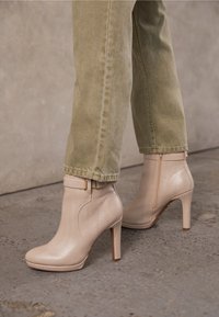 Bottines beiges à talons hauts avec fermetures éclair latérales et brides à boucle, portées avec un pantalon vert clair sur un sol en béton.
