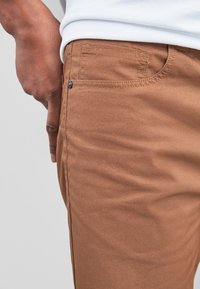 Pantalon en coton marron à la texture lisse, avec une poche latérale et un détail de bouton en métal, présenté avec une main reposant sur la hanche.