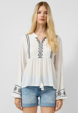 Femme aux longs cheveux blonds portant un chemisier blanc à manches longues avec une broderie noire et un short en jean bleu clair, debout sur un fond uni.