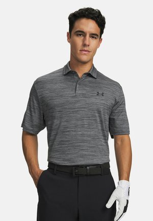 Uomo con polo grigia a motivo, pantaloni neri con cintura e guanto da golf bianco, in piedi con una mano in tasca su sfondo bianco.