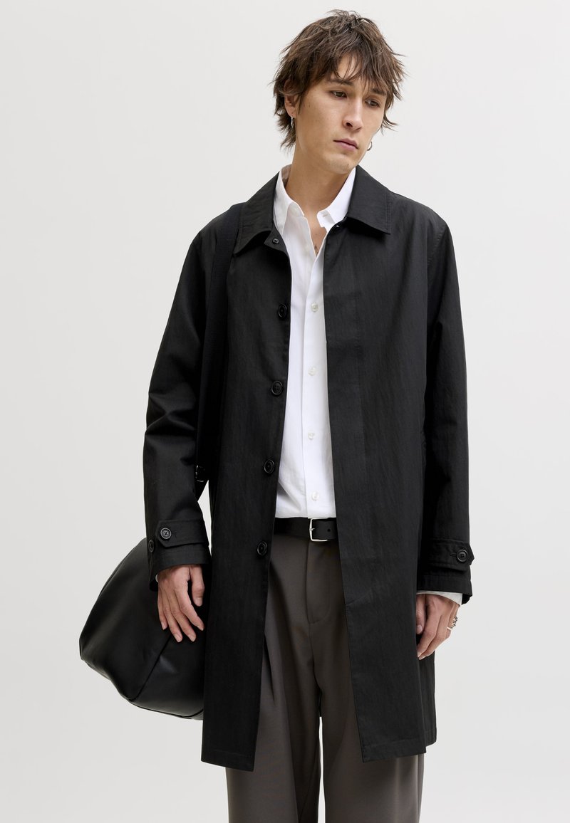 Junger Mann trägt einen schwarzen, geknöpften Mantel über einem weißen Hemd und grauen Hosen, hält eine große schwarze Tasche vor einfarbigem Hintergrund.