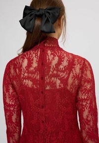 Červené čipkované šaty s jemnými kvetinovými vzormi, vysokým golierom a radom zodpovedajúcich gombíkov na chrbte. Vlasová stuha z čierneho saténu.