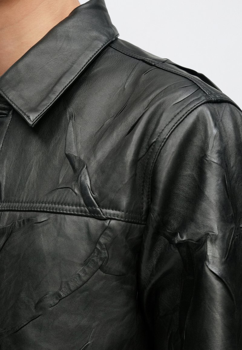 Veste en cuir noire avec une finition texturée, un col classique et des détails de coutures visibles le long de l'épaule et de la poitrine.