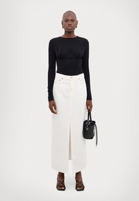 Zwarte lange mouw top met gatherdetails, gecombineerd met een witte high-waisted denim rok met een split aan de voorkant. Model houdt een kleine zwarte tas vast.