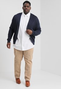 Marine cardigan over een witte blouse, gecombineerd met kaki broek en bruine schoenen. De stof lijkt glad met een klassieke pasvorm.