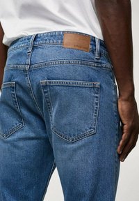 Modré džínové kalhoty s klasickým střihem, s hnědou koženou nášivkou s nápisem "MNG Denim" na mýtku a dvěma zadními kapsami.