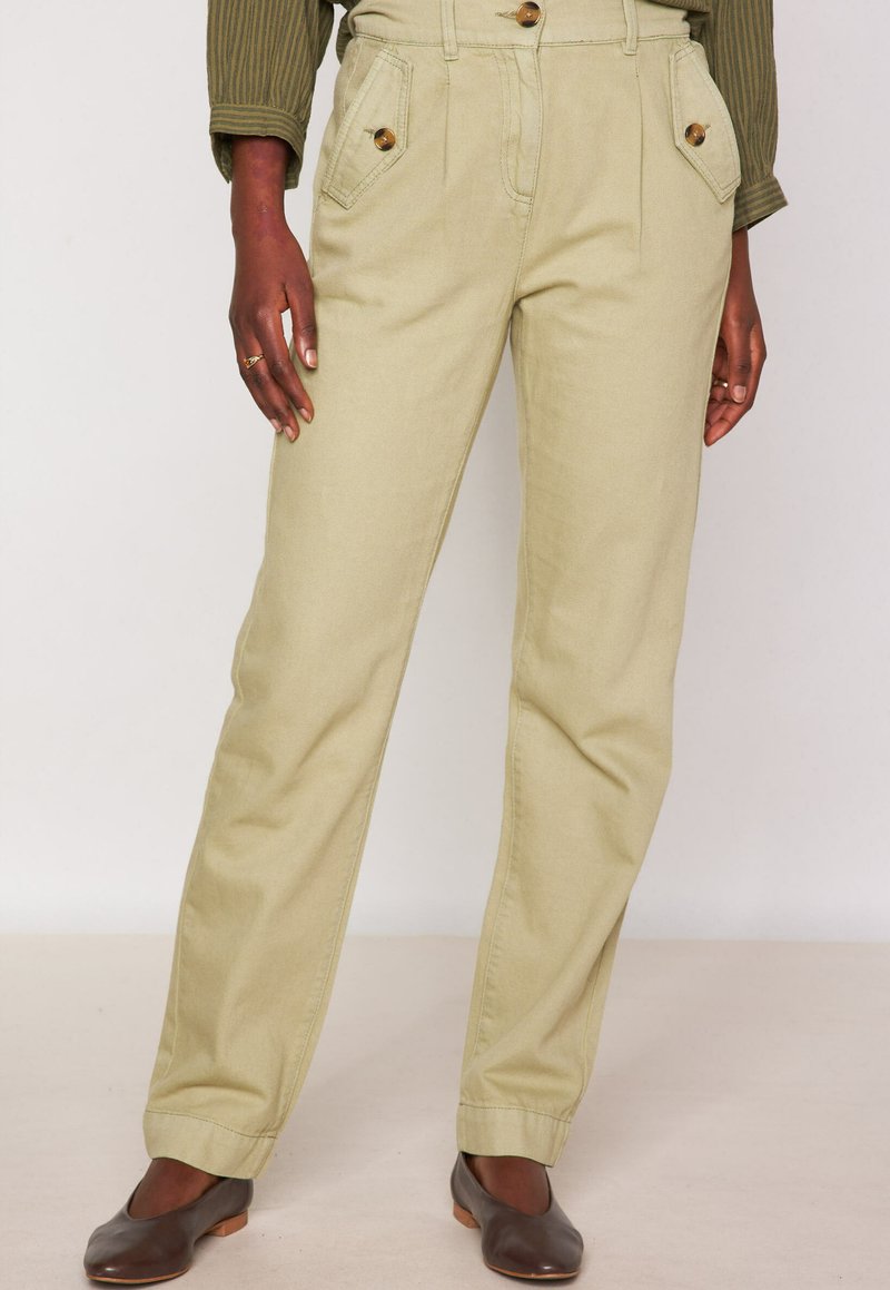 Persoon in beige hoge taille broek met knoopzakken aan de voorkant, gecombineerd met bruine ballerina's en een groene lange mouw top.