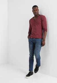 Langärmliges weinrot Henley-Shirt mit drei Knöpfen, kombiniert mit schmal geschnittenen blauen Jeans und schwarzen Sneakern. Einfaches Design, einfarbige Farben, minimalistische Muster.