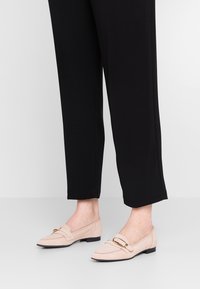 Lichtroze suède loafers met een platte zool, spitse neus en een gouden gesp accent. Draagt met zwarte tapered broeken.