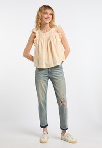 Top senza maniche color pesca chiaro con bordi arricciati, abbinato a jeans blu sbiaditi con ginocchio strappato e sneaker bianche.