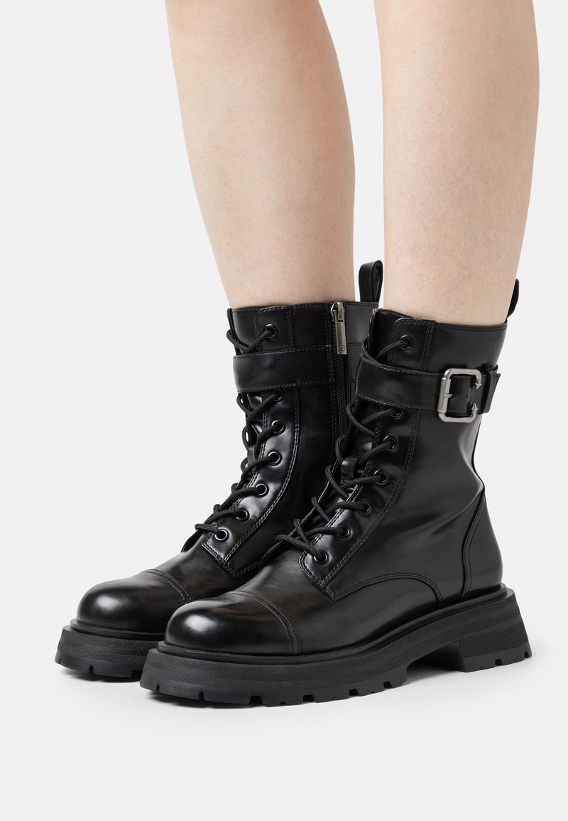 Botas de piel negra hasta el tobillo con puntera redondeada, parte delantera con cordones, cremallera lateral y una correa ancha con hebilla metálica. Suela de goma con textura.