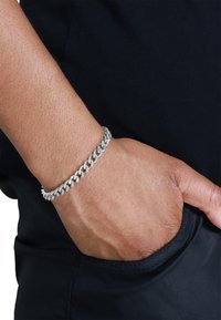 Bracelet en chaîne en argent sur un poignet, avec de gros maillons ovales entrelacés. La main repose sur une surface en tissu foncé.