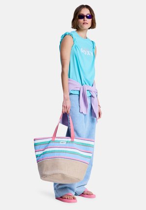 Jonge vrouw met een blauw topje met ruches aan de mouwen en zonnebril, met een gestreepte strandtas in haar hand en een lavendelkleurige trui om haar middel geknoopt.