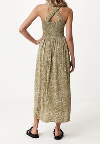 Vestido maxi verde con un cuerpo fruncido y tirantes entrecruzados. Presenta un diseño de hojas sobre un fondo claro y una silueta fluida.