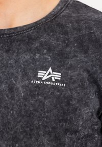 Osoba nosi crnu majicu s kiselim pranjem s bijelim Alpha Industries logotipom i natpisom na području grudiju.