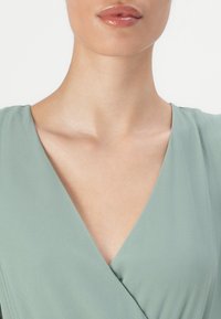 Ljusgrön omslagsklänning med V-ringning, texturerad tyg och mjuk drapering. Har släta, eleganta linjer och en figurnära silhuett.