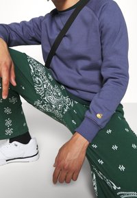 Sudadera morada con puños acanalados, combinada con pantalones verdes que presentan motivos de paisley blancos. Zapatillas blancas completan el atuendo.
