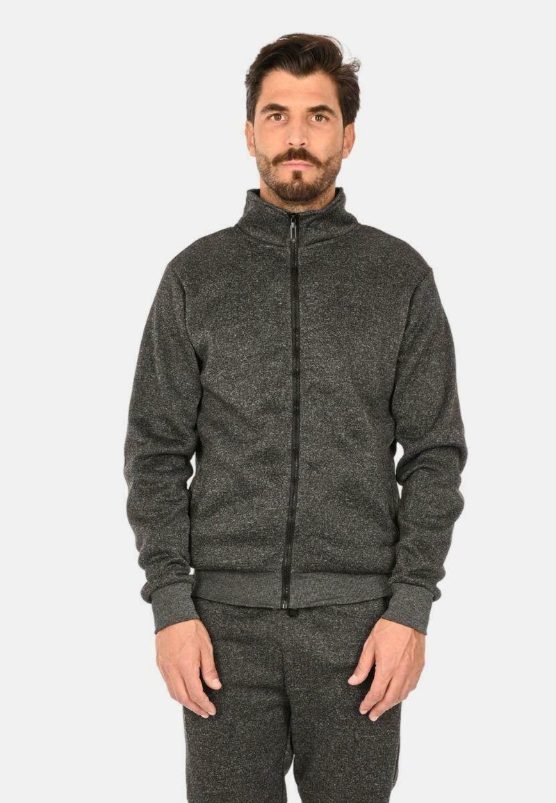 Giacca zip nera scura con colletto alto, tessuto testurizzato, polsini e orlo a costine, zip frontale e vestibilità rilassata. Abbinata a pantaloni coordinati.