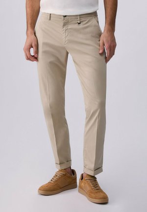 Uomo che indossa pantaloni beige slim fit abbinati a sneakers in pelle scamosciata marrone chiaro, in piedi contro uno sfondo chiaro e uniforme.
