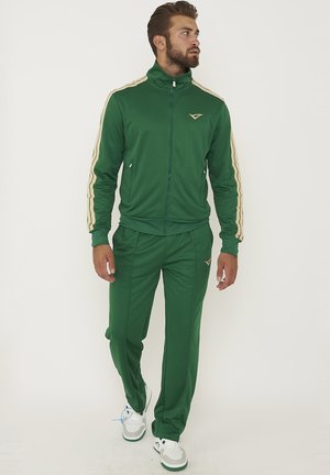Uomo che cammina indossando una tuta verde con strisce beige sulle maniche e sneaker bianche su sfondo bianco uniforme.