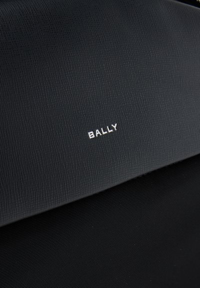 Κοντινή λήψη υφασμένου μαύρου δέρματος με μικρό ασημένιο λογότυπο "BALLY" ανάγλυφο στο κέντρο πάνω από ομαλό μαύρο ύφασμα.